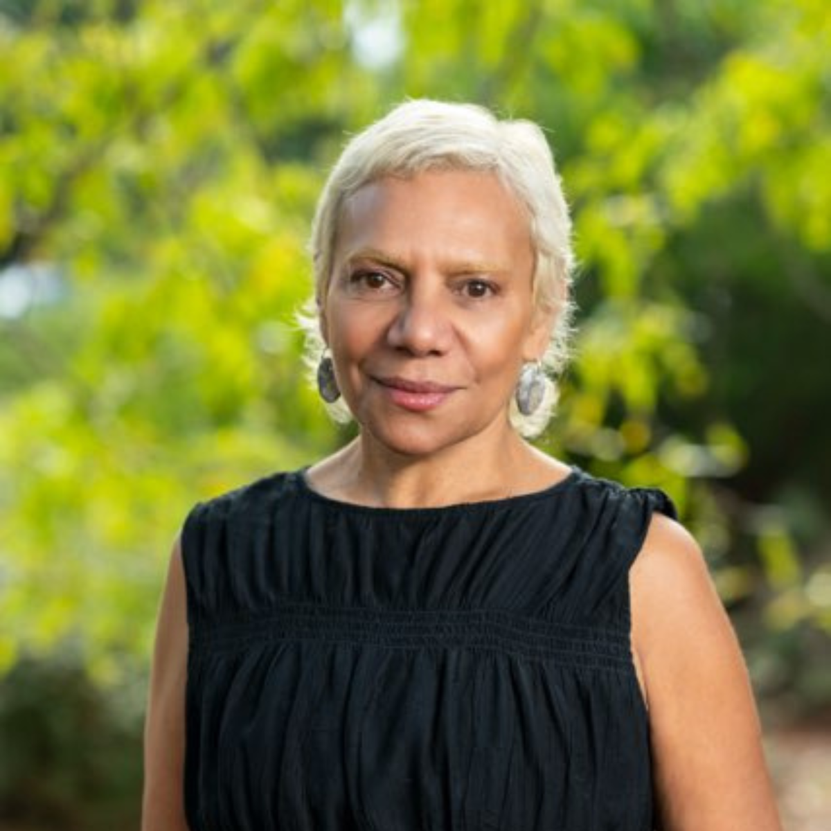 Portrait of a woman with short blonde hair outdoors, wearing a black sleeveless top and silver earrings, with greenery in the background